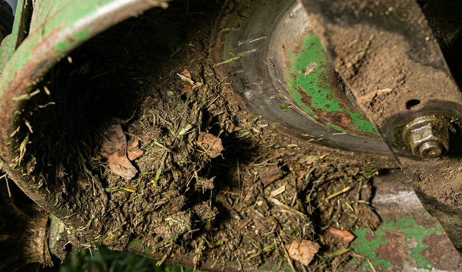 A close-up of a lawn mower's underside, heavily caked with wet grass clippings, mud, and debris, showing the rust and wear caused by buildup around the blade and deck.