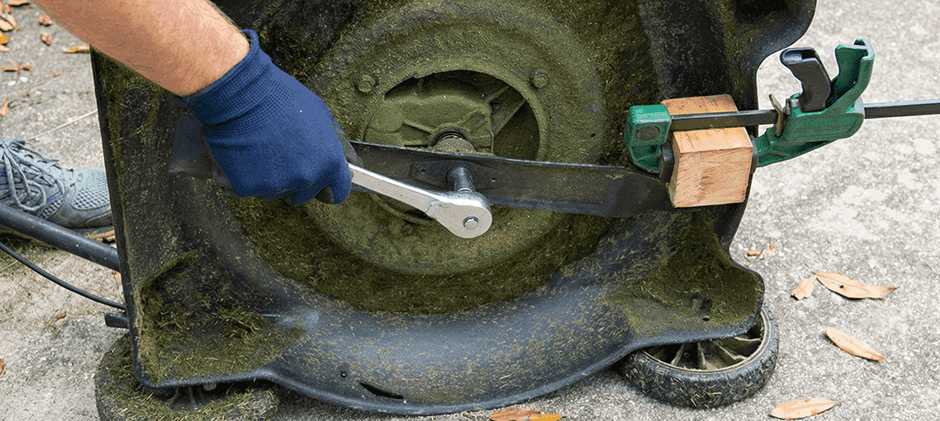 A socket wrench loosening a lawn mower blade bolt, with the blade held in place by a wood block and clamp.