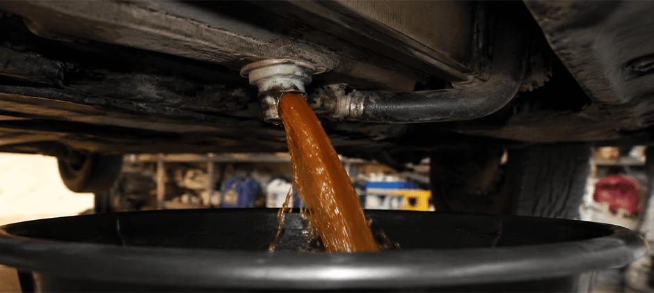 Old, rusty-brown coolant being drained from a vehicle's radiator into a black oil drain pan, representing the first step of a cooling system flush.