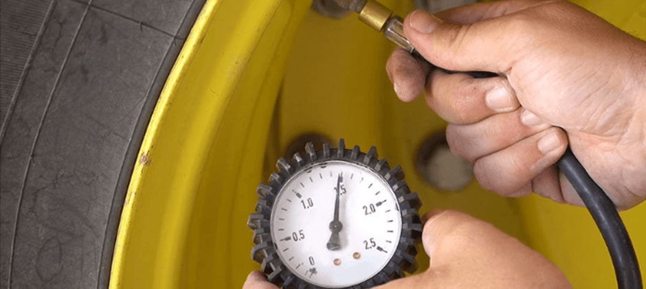 A person's hands using a dial pressure gauge to check the tire pressure on the valve stem of a large, yellow heavy equipment tire.