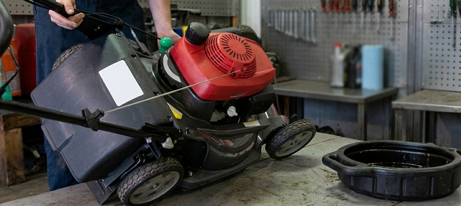 A push lawn mower is tilted on its side on a workbench to drain excess oil into a black collection pan below.