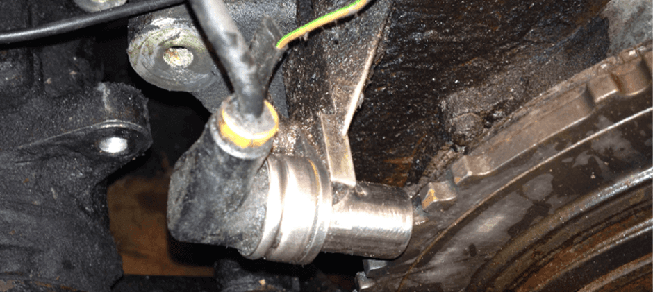 An old crankshaft position sensor installed in a dirty engine, pointing at the toothed reluctor wheel.