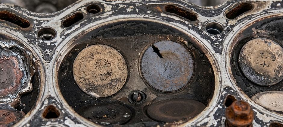 A close-up, top-down view of a dirty engine cylinder head, showing a visibly cracked and burnt exhaust valve.