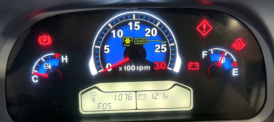 A close-up of a tractor's dashboard showing the RPM gauge, a low battery warning, and other system indicator lights on.