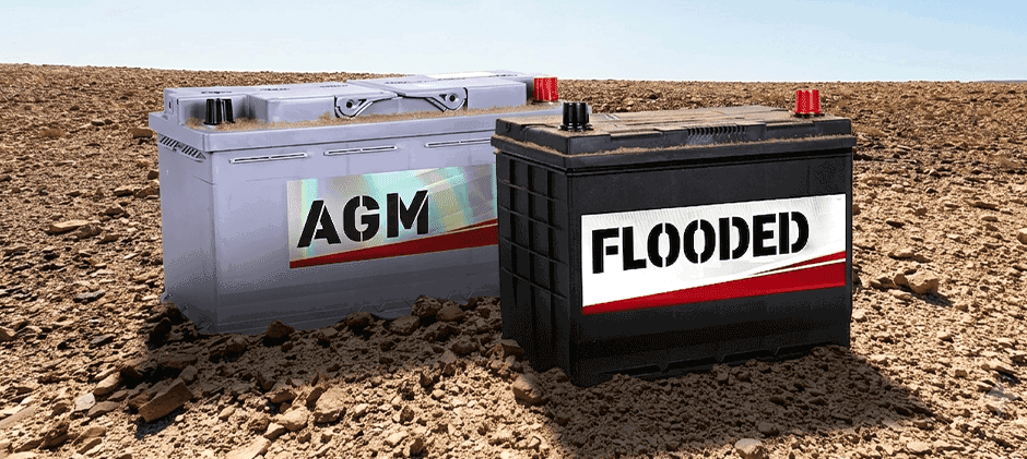 A gray AGM battery and a black flooded battery sit next to each other on dry, cracked earth, representing the two main battery options for farm equipment.
