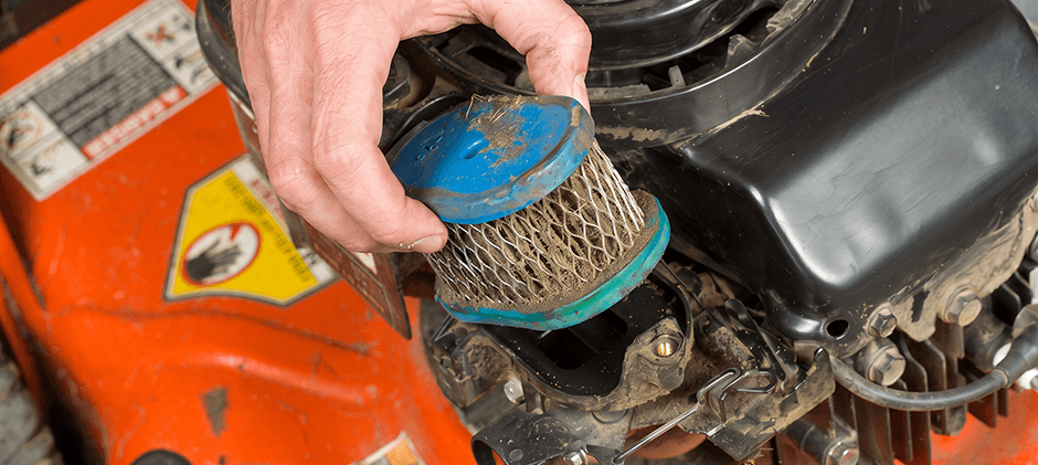 A hand lifting a clogged, dirty round air filter out of a lawn mower's engine housing.
