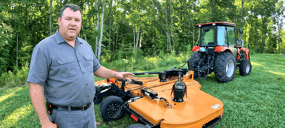 A man standing in a field next to an orange bush hog attached to a red tractor, pointing at the mower deck.