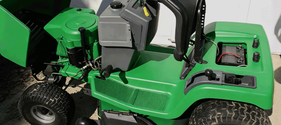An overhead view of a green riding mower with the hood open, showing the 12-volt battery, terminals, and engine wiring.