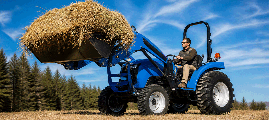 A man operating a blue LS tractor, using the front-end loader to lift a large round bale of hay in a field.