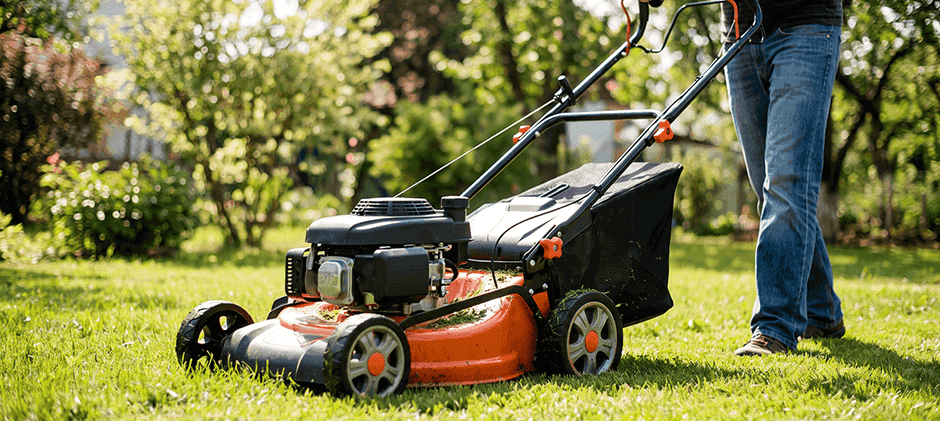 A person wearing jeans and walking shoes pushes an orange and black lawn mower across a sunny, green backyard lawn.