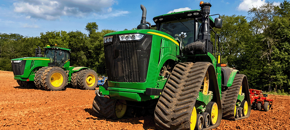 A large green tracked tractor pulling a tiller through a dry field, with another wheeled tractor visible in the background.