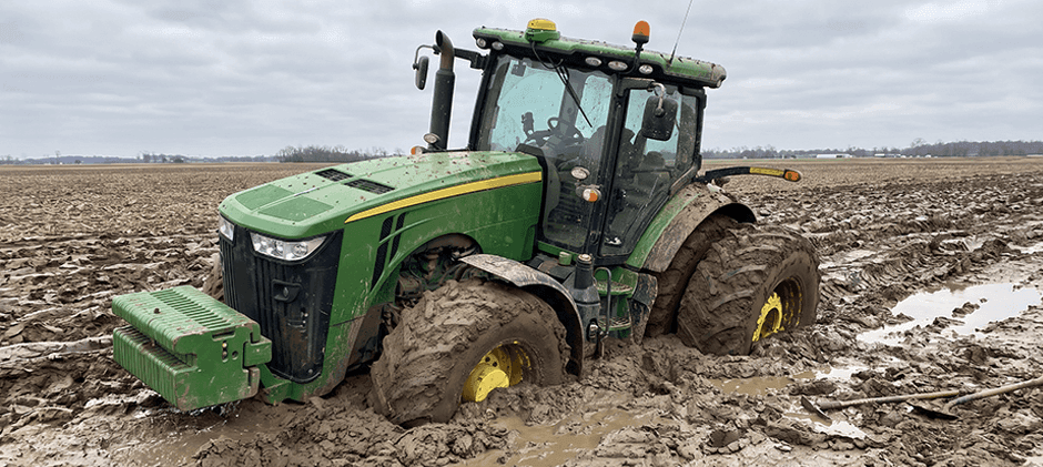 Large green tractor severely stuck in a muddy field with its axles buried deep in the ruts.