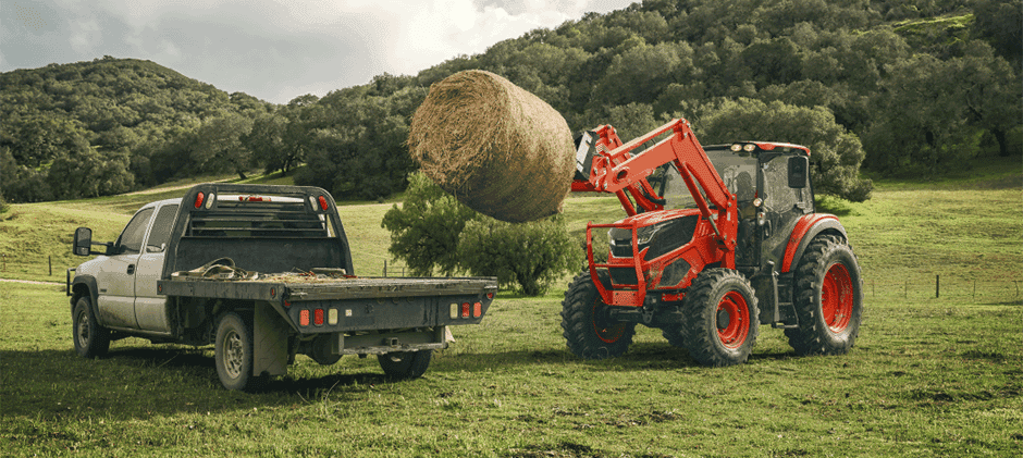An orange Kioti tractor with a front-end loader lifting a large round hay bale to place it on a white flatbed truck in a green pasture.