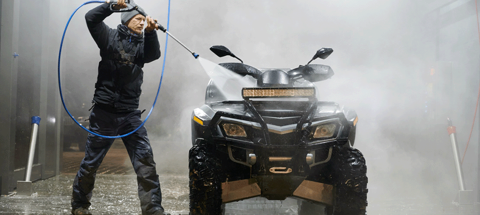 A man in a winter jacket using a pressure washer on a black ATV in a commercial wash bay, using a wide spray pattern to protect the machine.