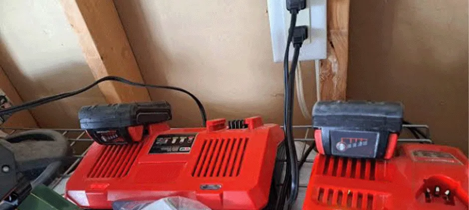 Dos cargadores de baterías Milwaukee conectados a una toma de corriente y colocados sobre una estantería metálica en un taller, representando una configuración habitual de carga.