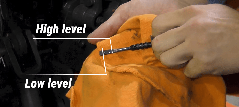 A person holds a clean orange rag while displaying a Kubota tractor's hydraulic fluid dipstick, with text pointing to the high and low level marks.