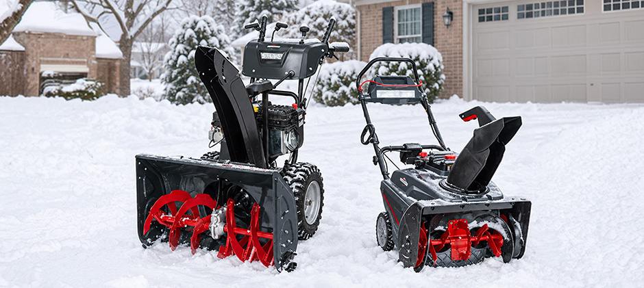 A single-stage and a two-stage snow blower parked side-by-side on a snowy residential driveway.